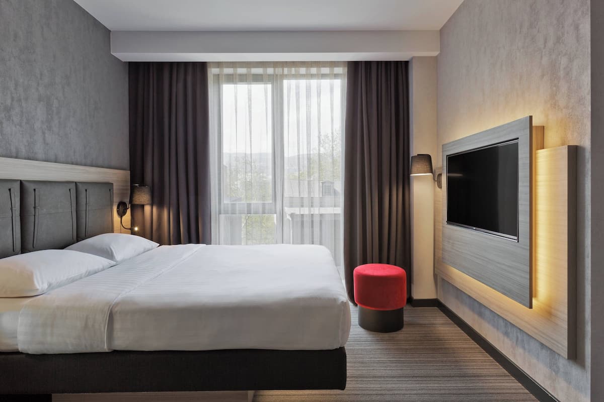 A modern hotel room with a large bed, a flat-screen TV mounted on the wall, and a view of the outdoors through large windows.