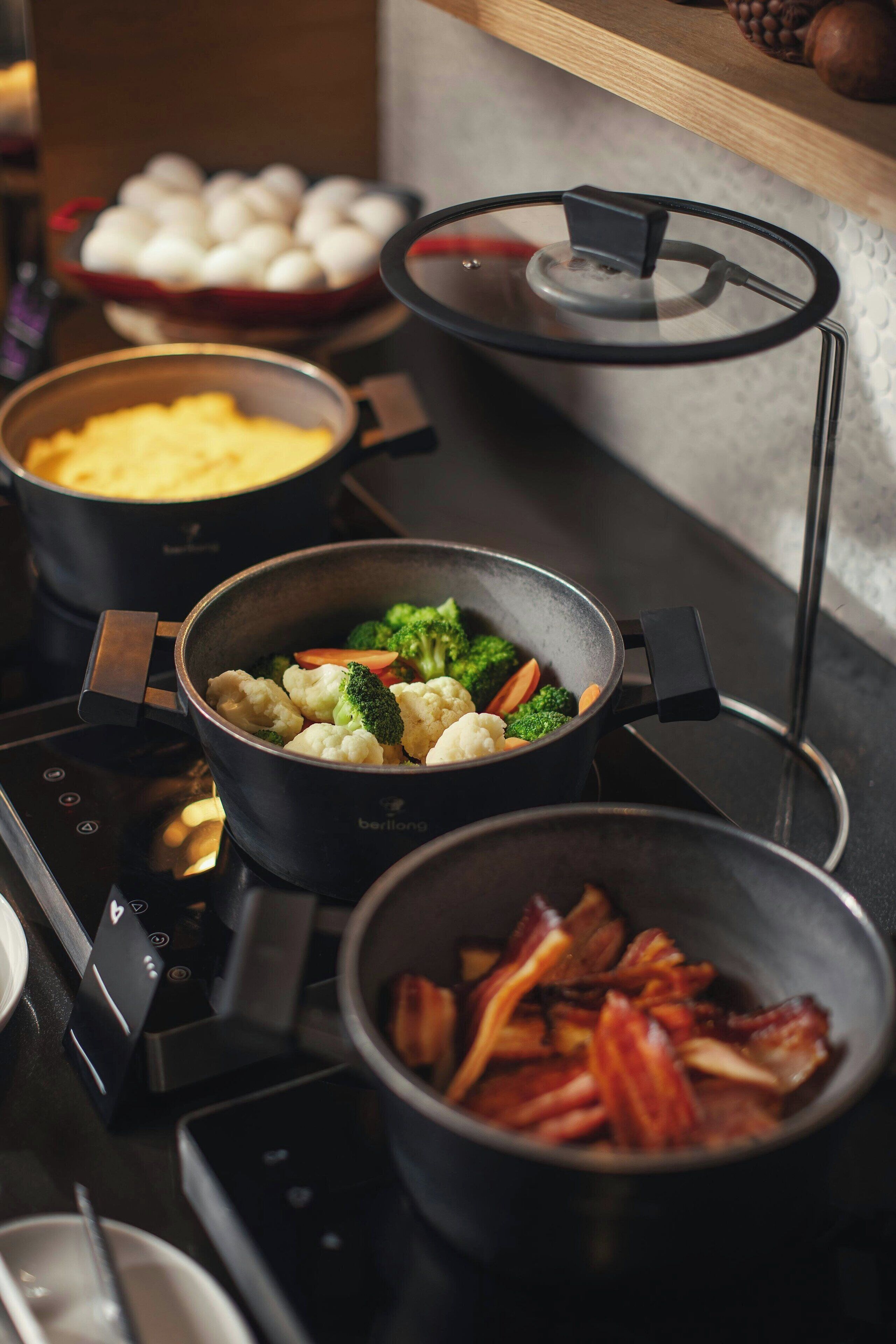 Et køkken med flere gryder på en induktionskogetop, herunder en med broccoli og blomkål, en anden med bacon, og en tredje med æg.