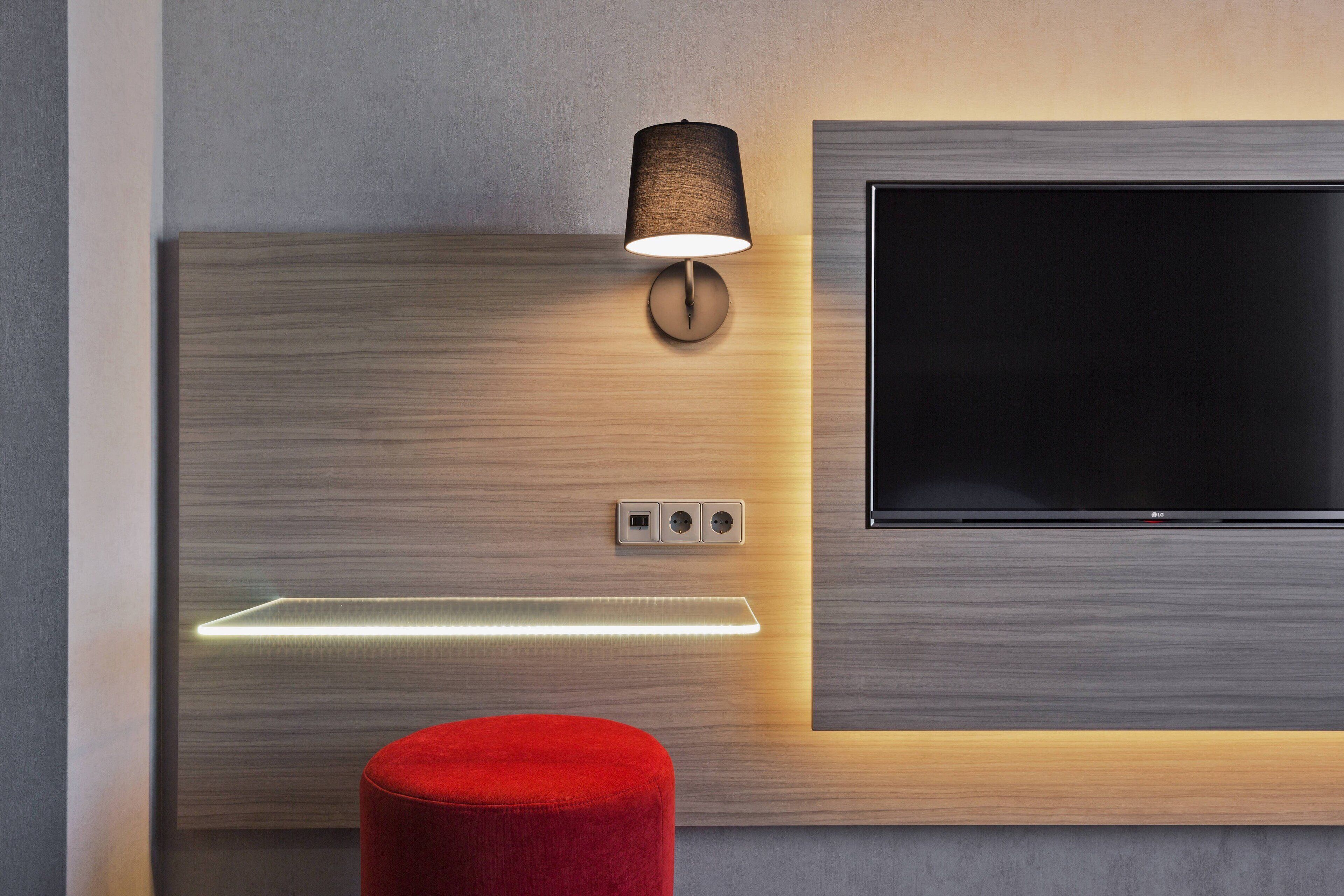 Una habitación moderna con una televisión montada en la pared, una lámpara de pared y un reposapiés rojo.
