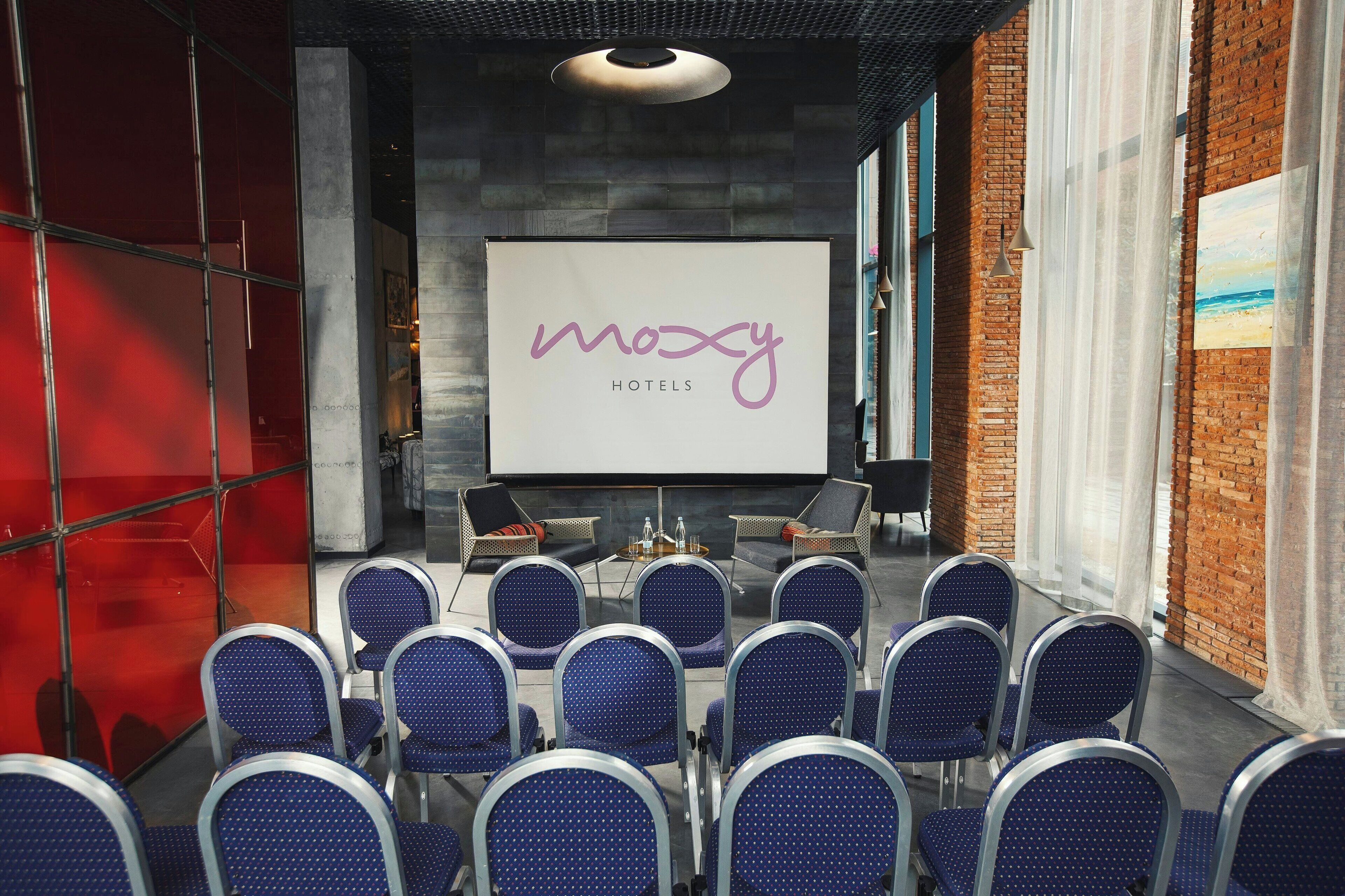 Un salón de conferencias con una pantalla grande mostrando la marca 'moosy hotels', filas de sillas azules y un fondo de pared de ladrillo.
