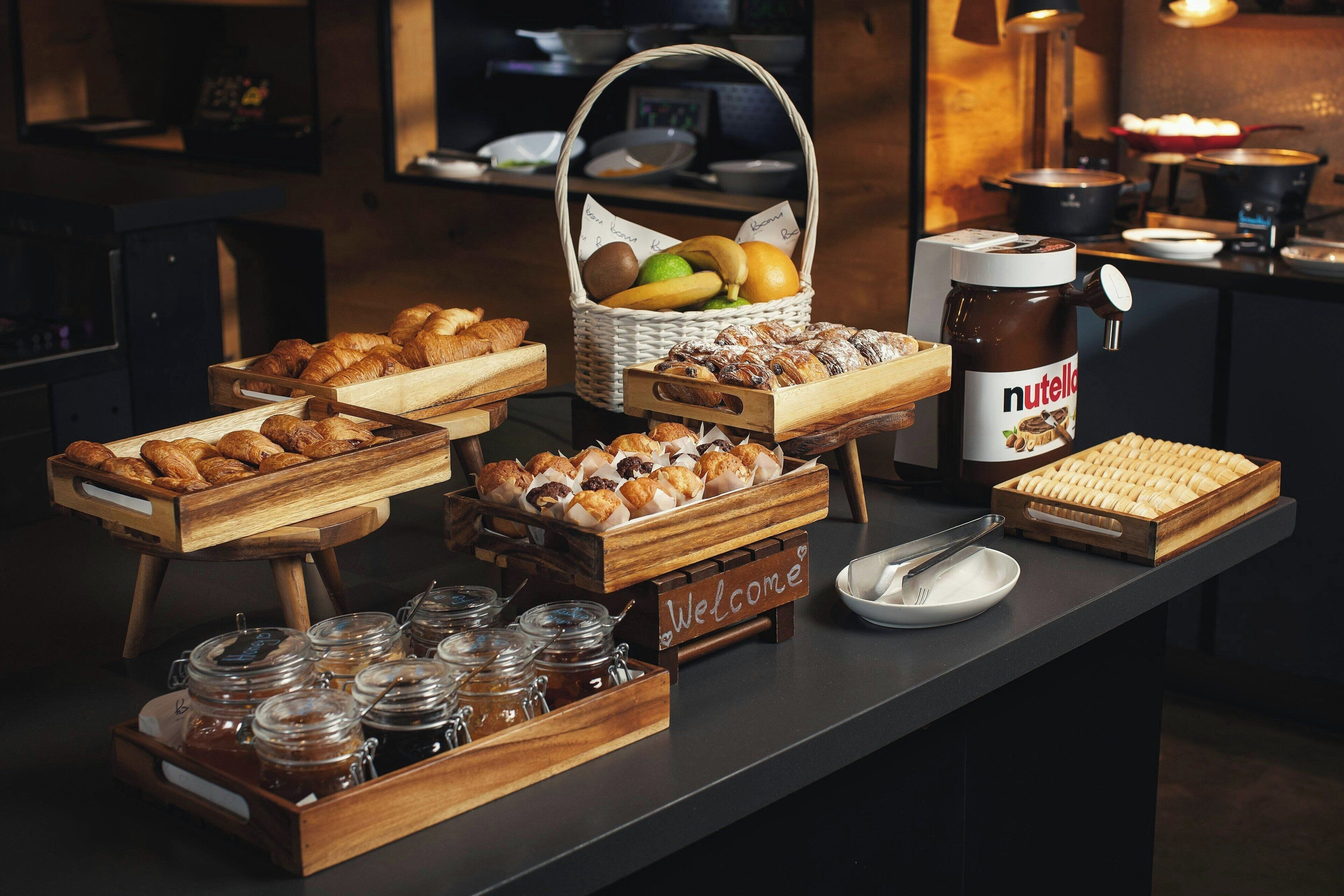 Una mesa de buffet con diversos pasteles, una cesta de frutas y un tarro de Nutella.