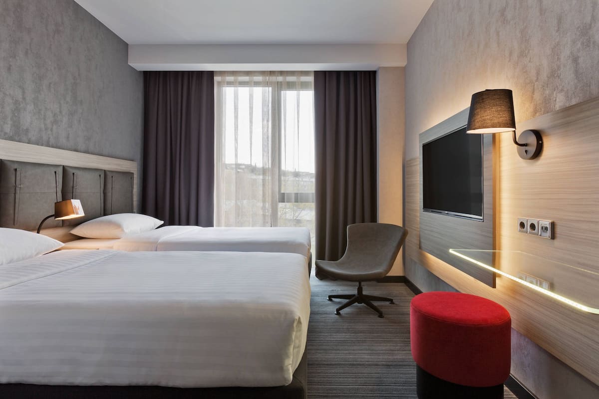 A hotel room with two beds, a TV, a chair, and a red ottoman.