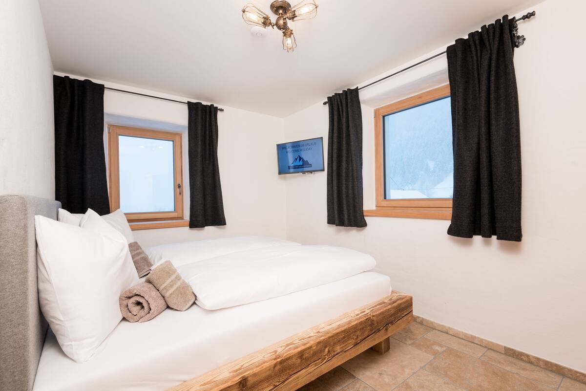 Quarto com estrutura de cama de madeira, roupa de cama branca, duas janelas com cortinas pretas, luminária no teto e um letreiro de boas-vindas na parede.