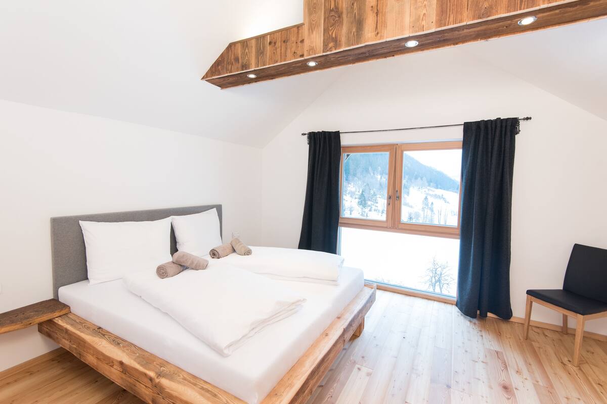 Quarto com estrutura de cama de madeira, cabeceira cinza e uma janela grande com cortinas.