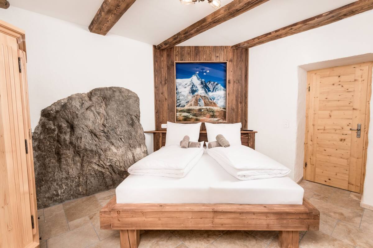 Uma cama de madeira com roupa de cama e toalhas brancas, uma grande rocha e uma pintura de montanha na parede.