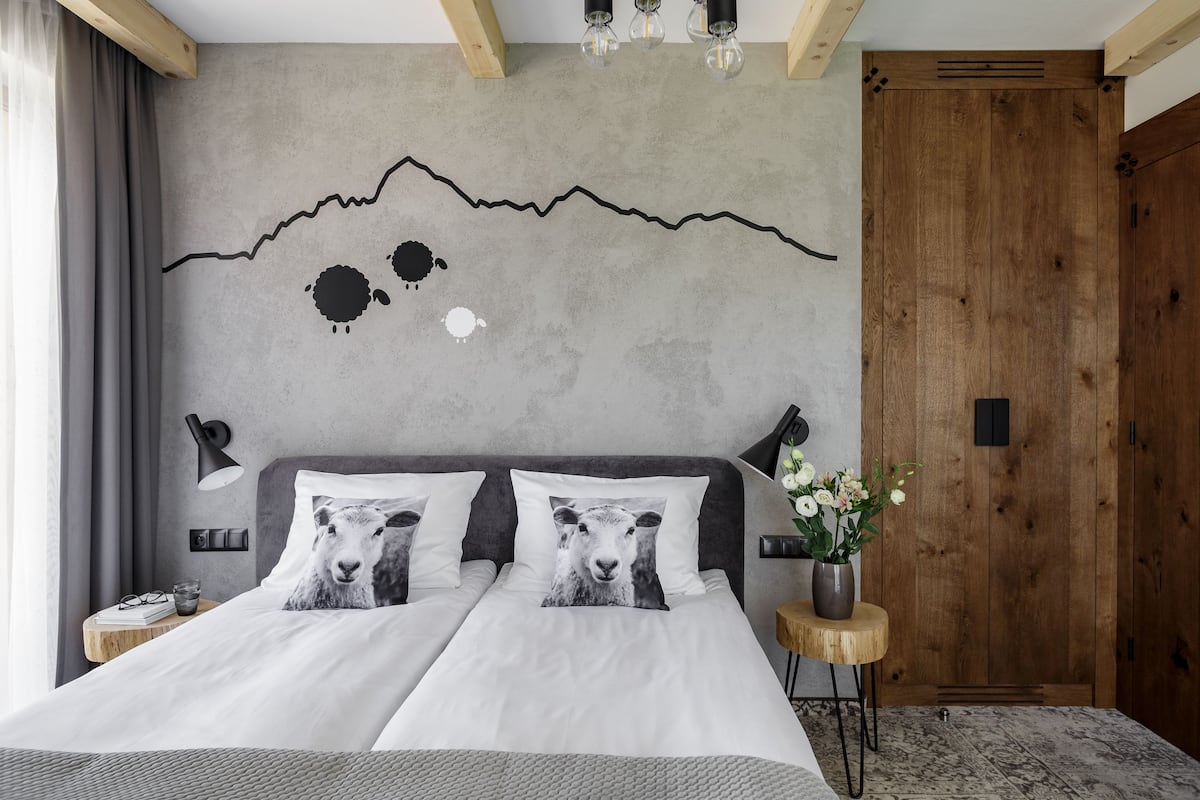 A bedroom with a bed, two pillows, a bedside table, a nightstand, a wall-mounted lamp, a wooden door, and a small wooden stool with a vase of flowers.