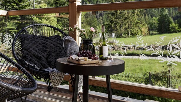 En træterrasse med et rundt bord, en fletstol og udsigt over et bjerglandskab.