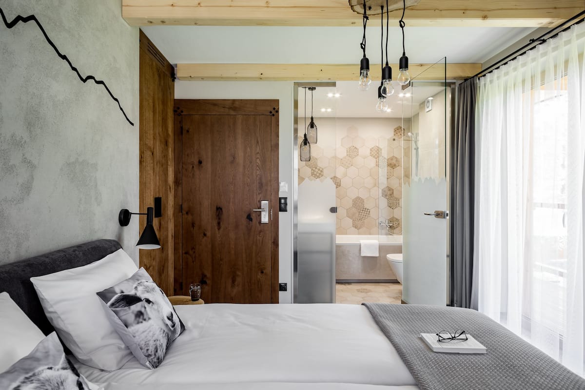 A modern bedroom with a wooden door, a bed with white bedding, a bedside table with glasses, and a bathroom visible through a glass door.