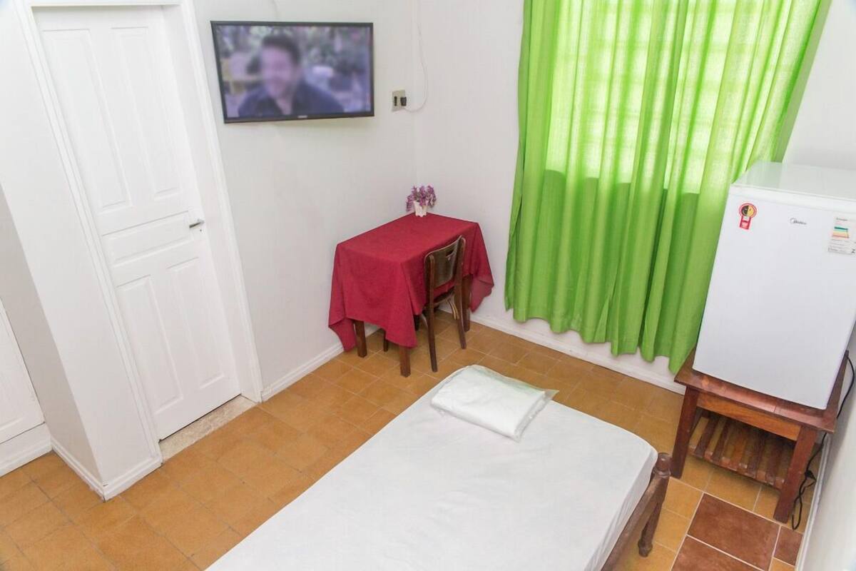 Una habitación con cama, una mesita con mantel rojo, un televisor en la pared y una cortina verde.