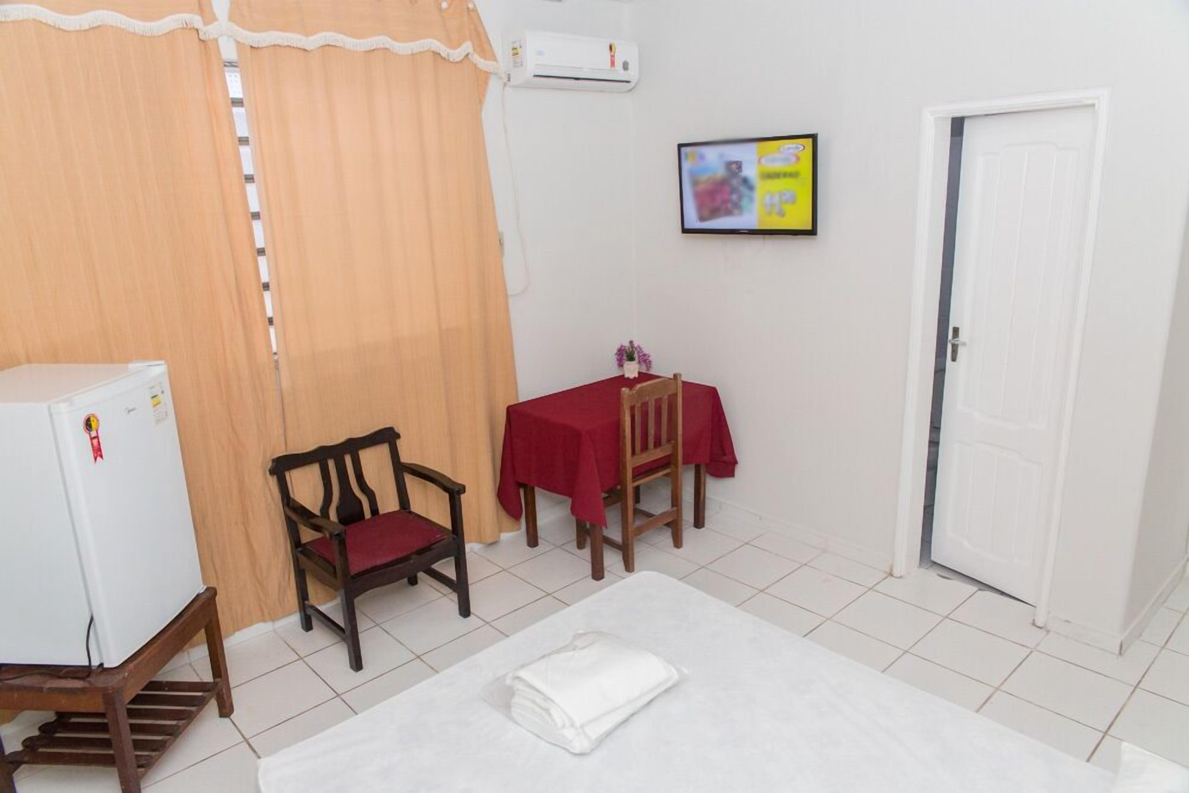 A small room with a bed, a table with a red tablecloth, a chair, a white refrigerator, and a door.