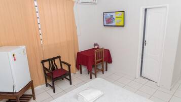 A small room with a bed, a table with a red tablecloth, a chair, a white refrigerator, and a door.