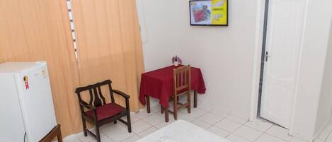 A small room with a bed, a table with a red tablecloth, a chair, a white refrigerator, and a door.