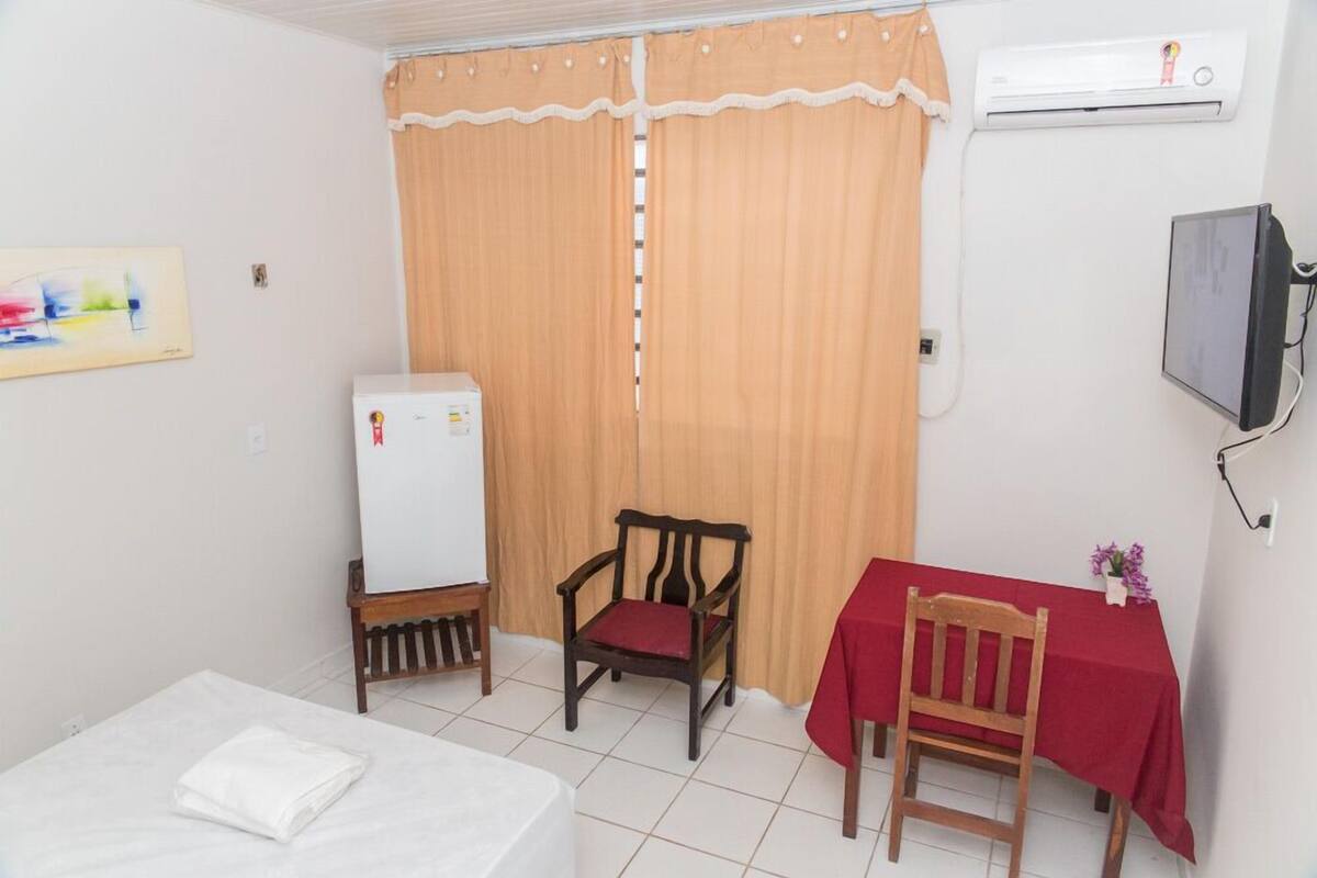 Una habitación pequeña con una cama, una mesa con mantel rojo, una silla, una silla de madera, una mesa pequeña con un objeto blanco, un televisor montado en la pared y una ventana con cortinas.