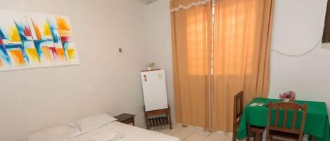 A room with a bed, a table with a green tablecloth, a refrigerator, and a window with curtains.