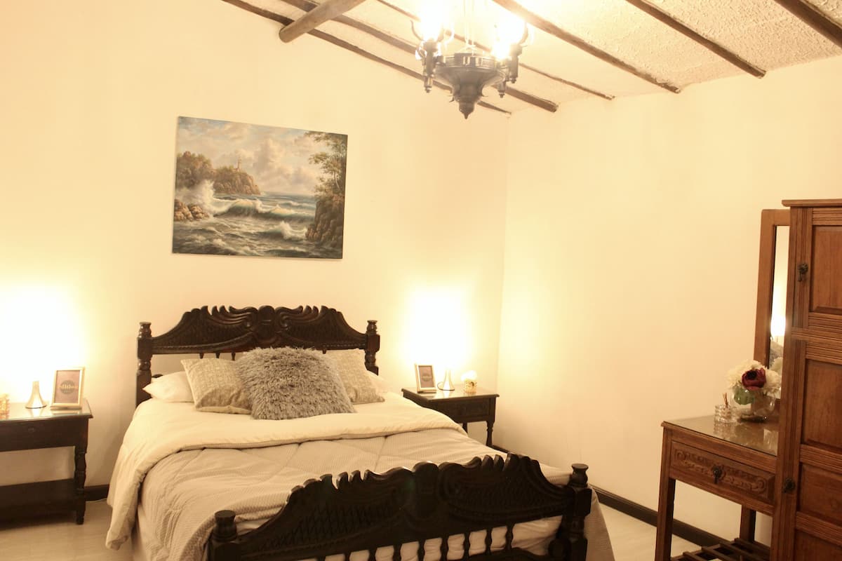 A bedroom with a wooden bed, two bedside tables, a nightstand, a dresser, a painting on the wall, and a chandelier.