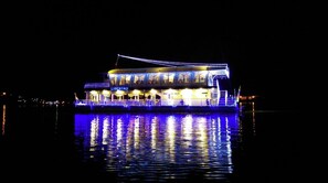 Un barco grande e iluminado con varias cubiertas y una bandera, flotando sobre el agua de noche.