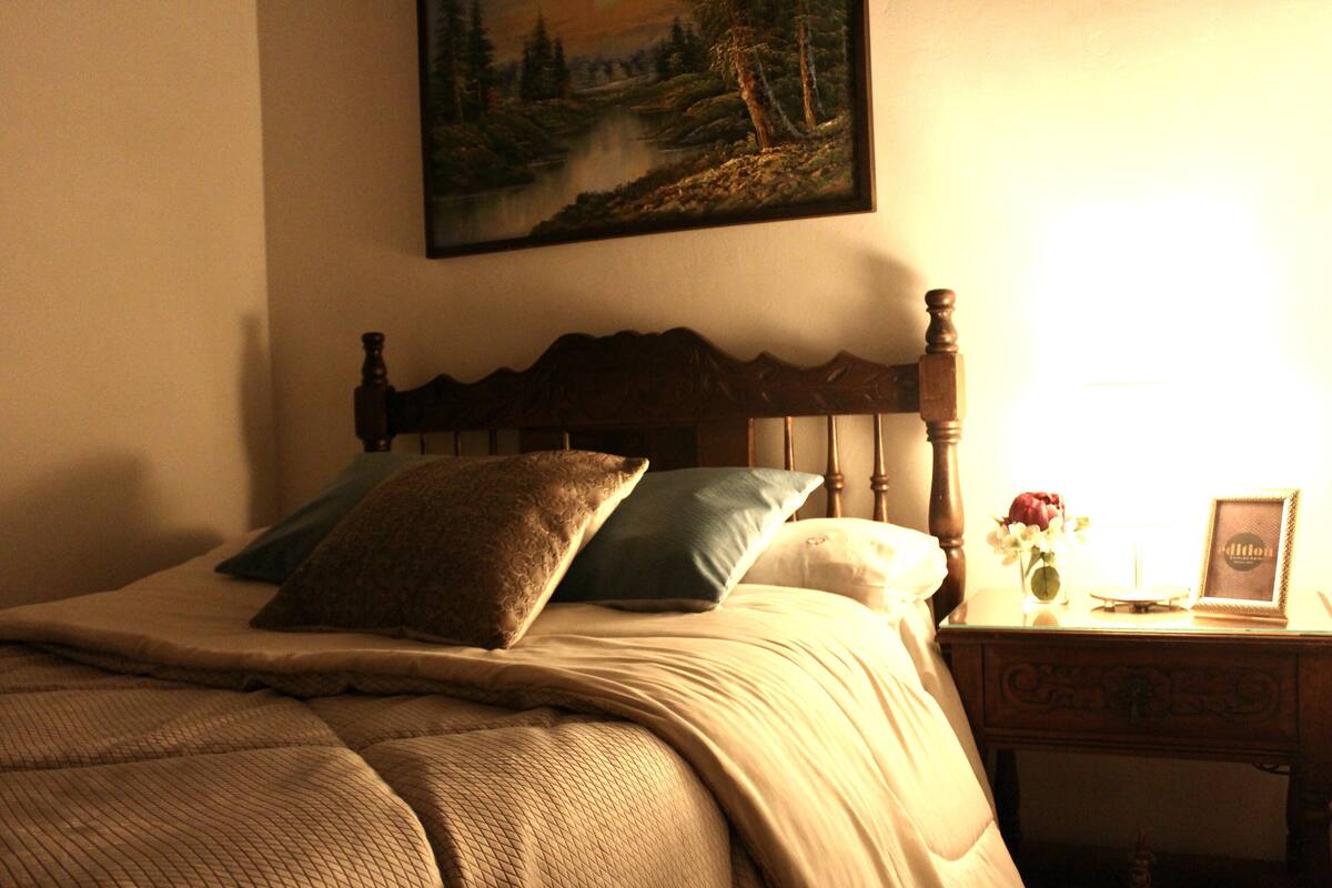 A bed with a wooden headboard, a bedside table with a vase and a picture frame, and a painting on the wall.