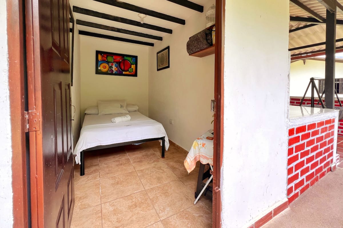 Un dormitorio con una cama, una mesita con mantel, una pared con un cuadro colorido y una pared de ladrillo rojo.