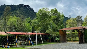 A playground with swings and slides, surrounded by a grassy area and a forested hill in the background. - Paraiso Cocora (Salento)