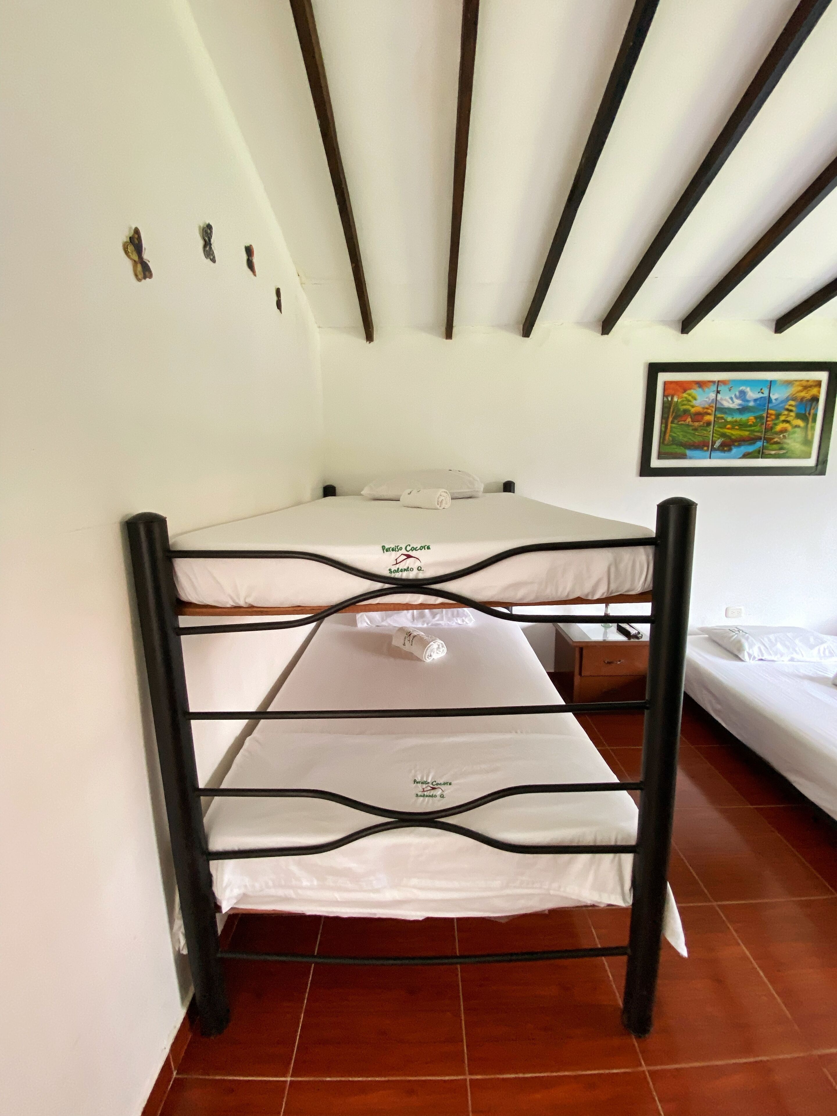 Een stapelbed met een witte matras, gelabeld 'Privado Cocote', en een enkel bed met een witte matras in een kamer met houten balken in het plafond en een schilderij aan de muur.