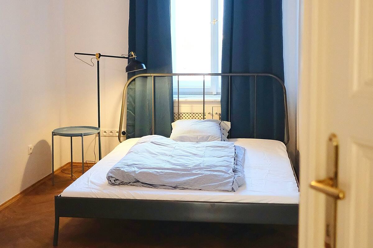 A bedroom with a bed, a standing lamp, a small table, and a window with curtains.
