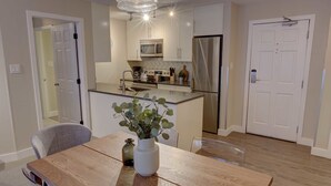 Une cuisine moderne avec des meubles blancs, des appareils en acier inoxydable et une table à manger en bois agrémentée d’un vase de plantes vertes.