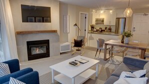 Un salon avec une cheminée, un coin repas avec une table et des chaises, et une cuisine à l’arrière-plan.