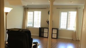 A spacious living room with hardwood flooring, a black sofa, a small table, and a lamp. There are two windows with curtains, a clock on the wall, and a ceiling light. - Walkout Ravine, Cottage feeling basement (Richmond Hill)