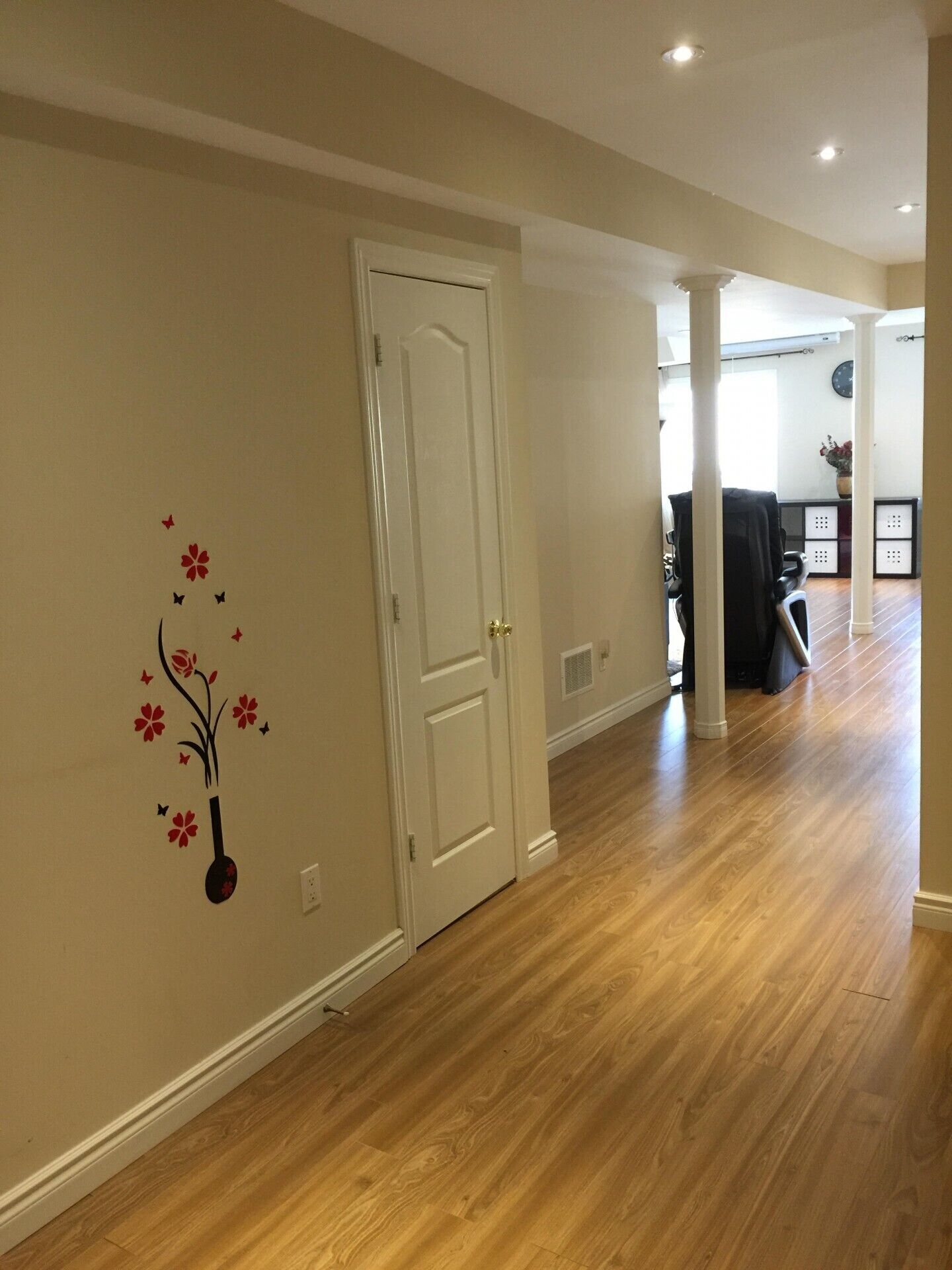 Un couloir avec un plancher en bois, une porte blanche et un motif décoratif représentant un vase de fleurs sur le mur.