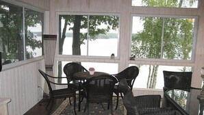 A covered patio with wicker furniture, a patterned rug, and large windows offering a view of trees and water. - Lake Muskoka in Muskoka Bay (Gravenhurst)