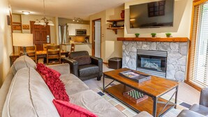 A living room with a sofa, coffee table, and fireplace. There is a television mounted above the fireplace and a kitchen in the background. - Aspens #444 Prime Ski-in Ski-out Location! Pool, Hot tubs, BBQ, sleeps 6 (Whistler)