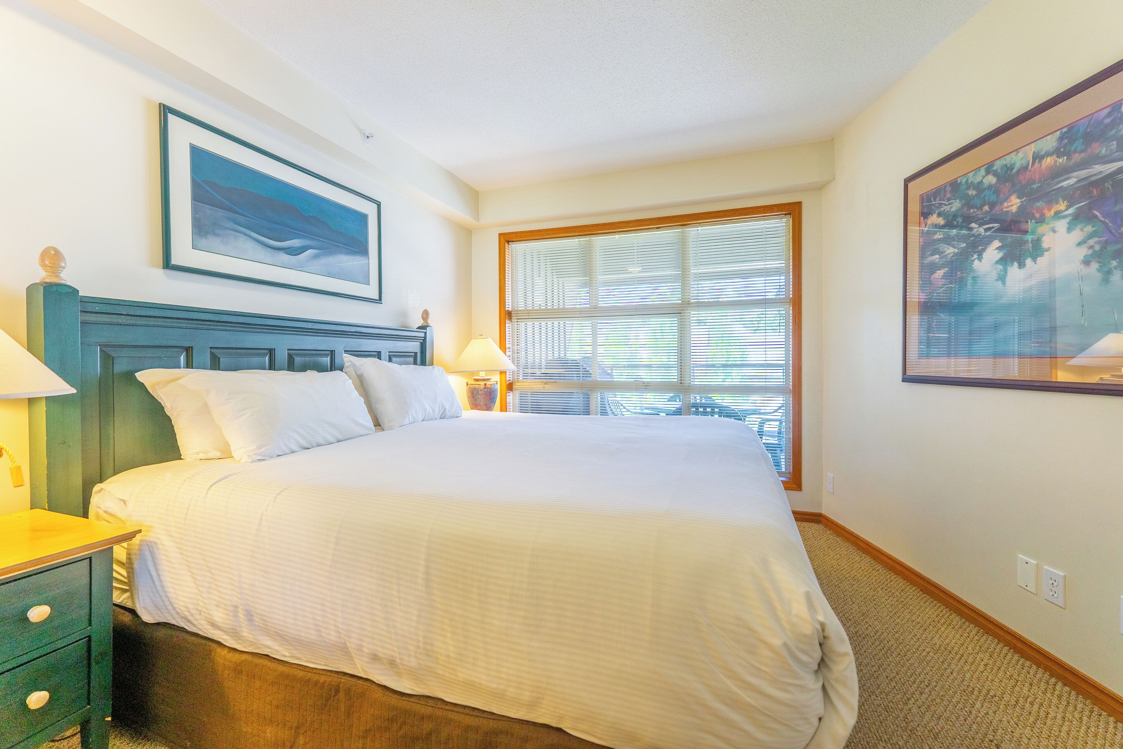 A neatly made bed with white linens, a wooden headboard, and a nightstand with a lamp. There is a window with blinds, a framed painting on the wall, and a carpeted floor.