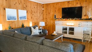 A cozy living room with a large sectional sofa, a flat-screen TV mounted on the wall, and a fireplace with a mantel decorated with holiday lights. - Family Friendly Stoney Lake Cottage (Lakefield)