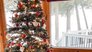 A Christmas tree decorated with red and white ornaments, set in a room with a wooden floor and large windows overlooking a deck. - Family Friendly Stoney Lake Cottage (Lakefield)