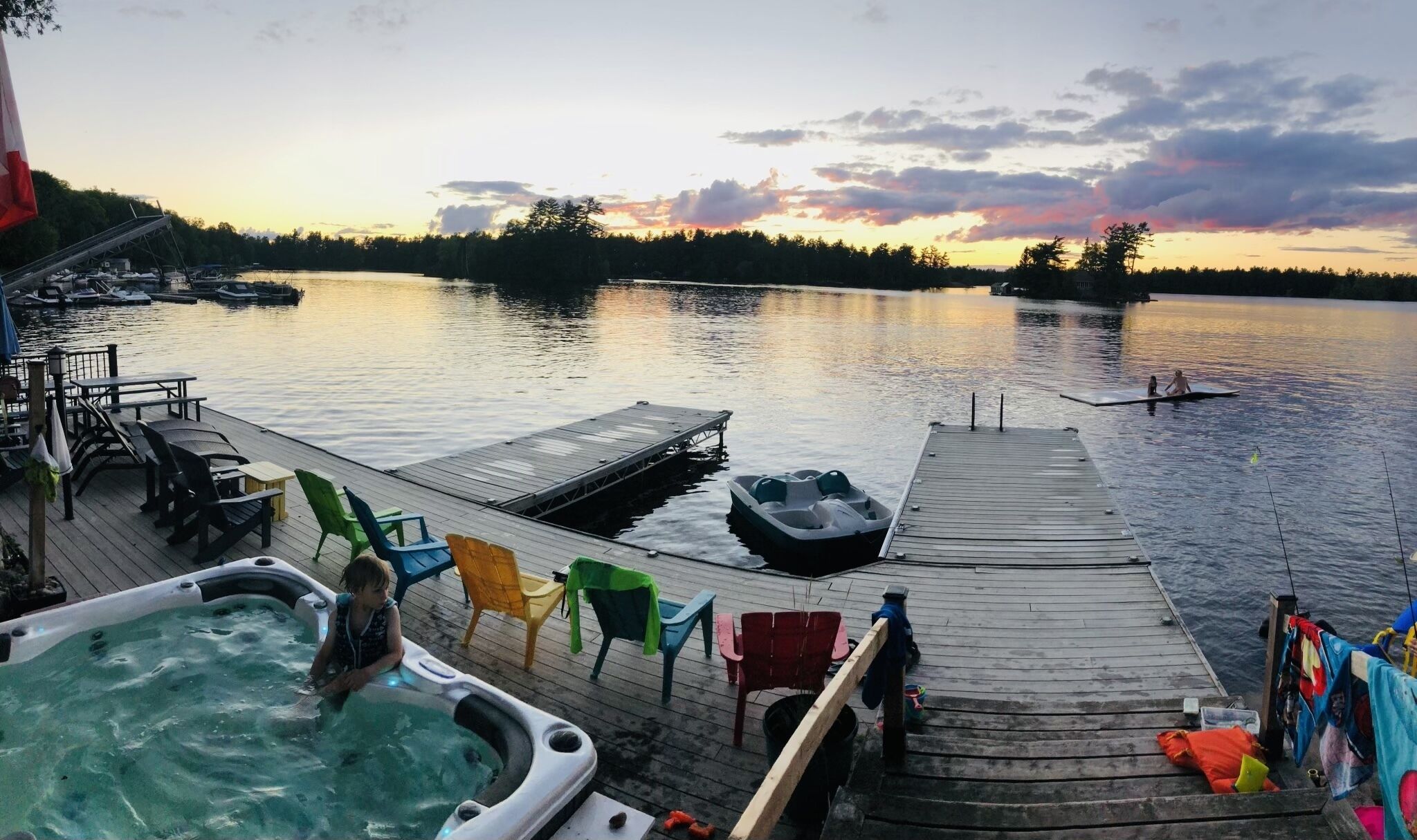 A wooden dock with hot tubs and colorful chairs, overlooking a calm lake at sunset.