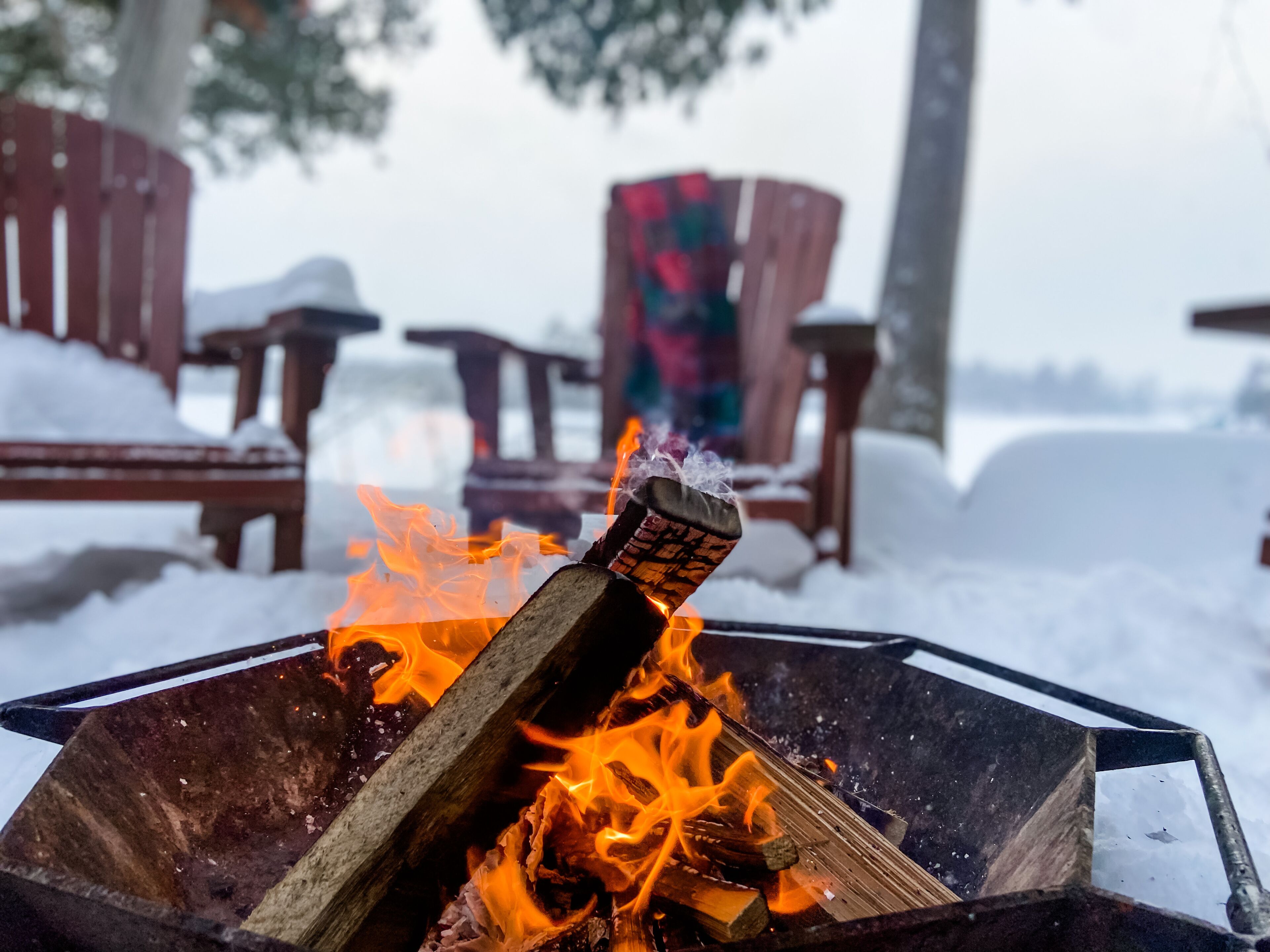 Een vuurkorf met brandende blokken hout in een metalen bak, omringd door besneeuwde stoelen.