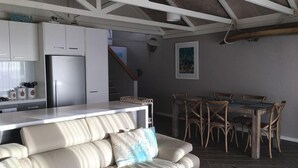 A modern kitchen with white cabinets and a stainless steel refrigerator. There is a dining area with wooden chairs and a table. A beige sofa with decorative pillows is in the foreground.