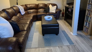 A living room with a brown leather sectional sofa, a black ottoman, a floor lamp, a painting, a bookshelf, and a television.
