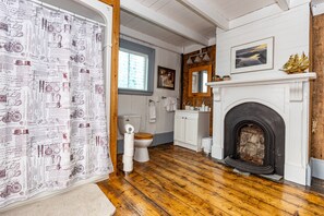 A bathroom with a wooden floor, a white toilet, a white sink, a mirror, and a fireplace. - Traditional cottage in historic trinity (Trinity, Trinity Bay)
