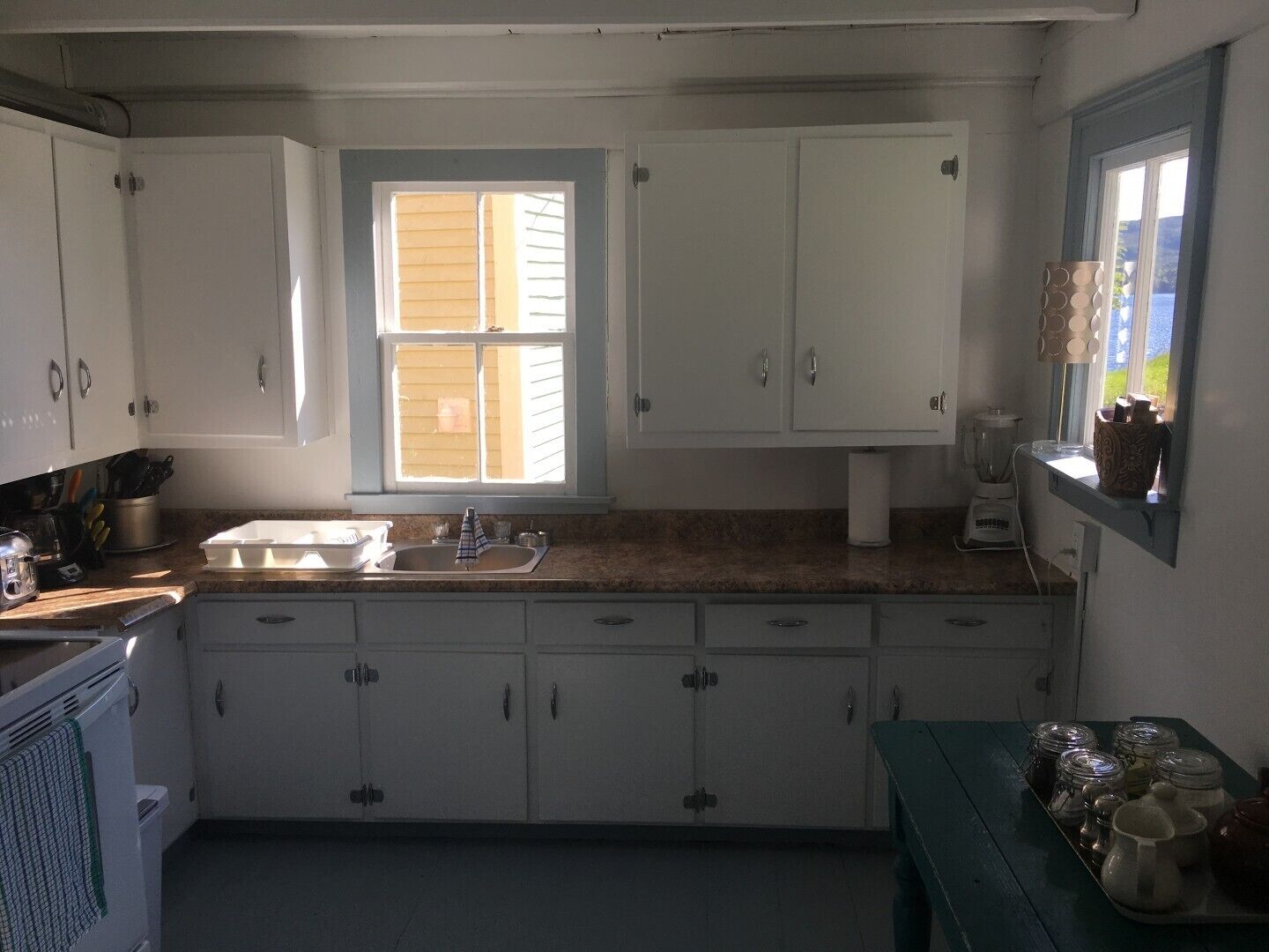 A kitchen with white cabinets, a window with blinds, a sink, a countertop with a blender, and a table with jars.
