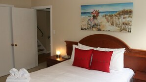Habitación de hotel con cama, un cuadro de una escena de playa, una mesita de noche con lámpara y una puerta que da a una escalera.