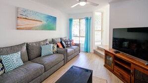 Un salon avec un canapé gris, des coussins colorés, une table basse noire, un meuble TV en bois et un tableau accroché au mur.