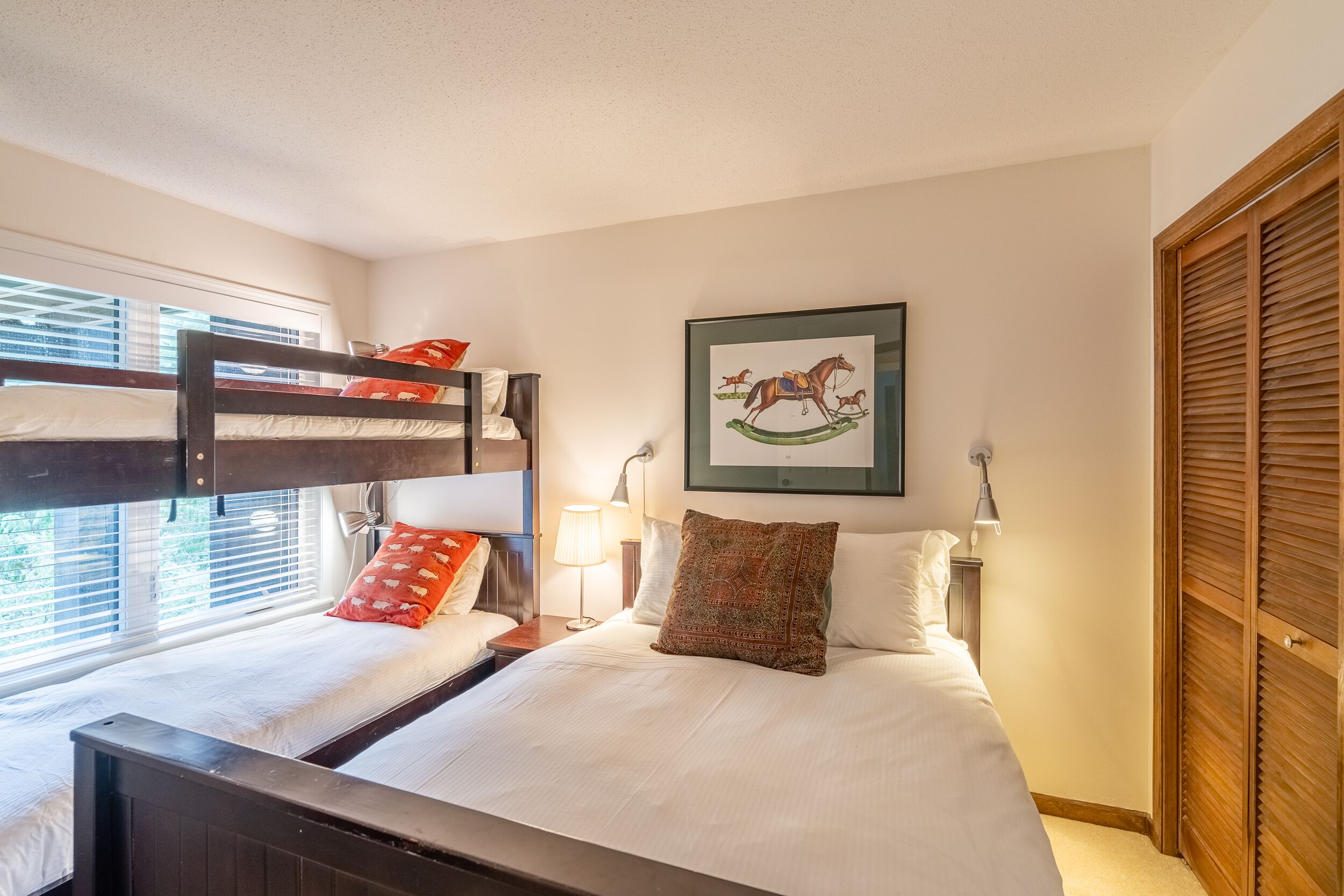 Une chambre à coucher avec un lit superposé, un lit simple, un tableau encadré représentant un cheval, une lampe de chevet et une armoire en bois.