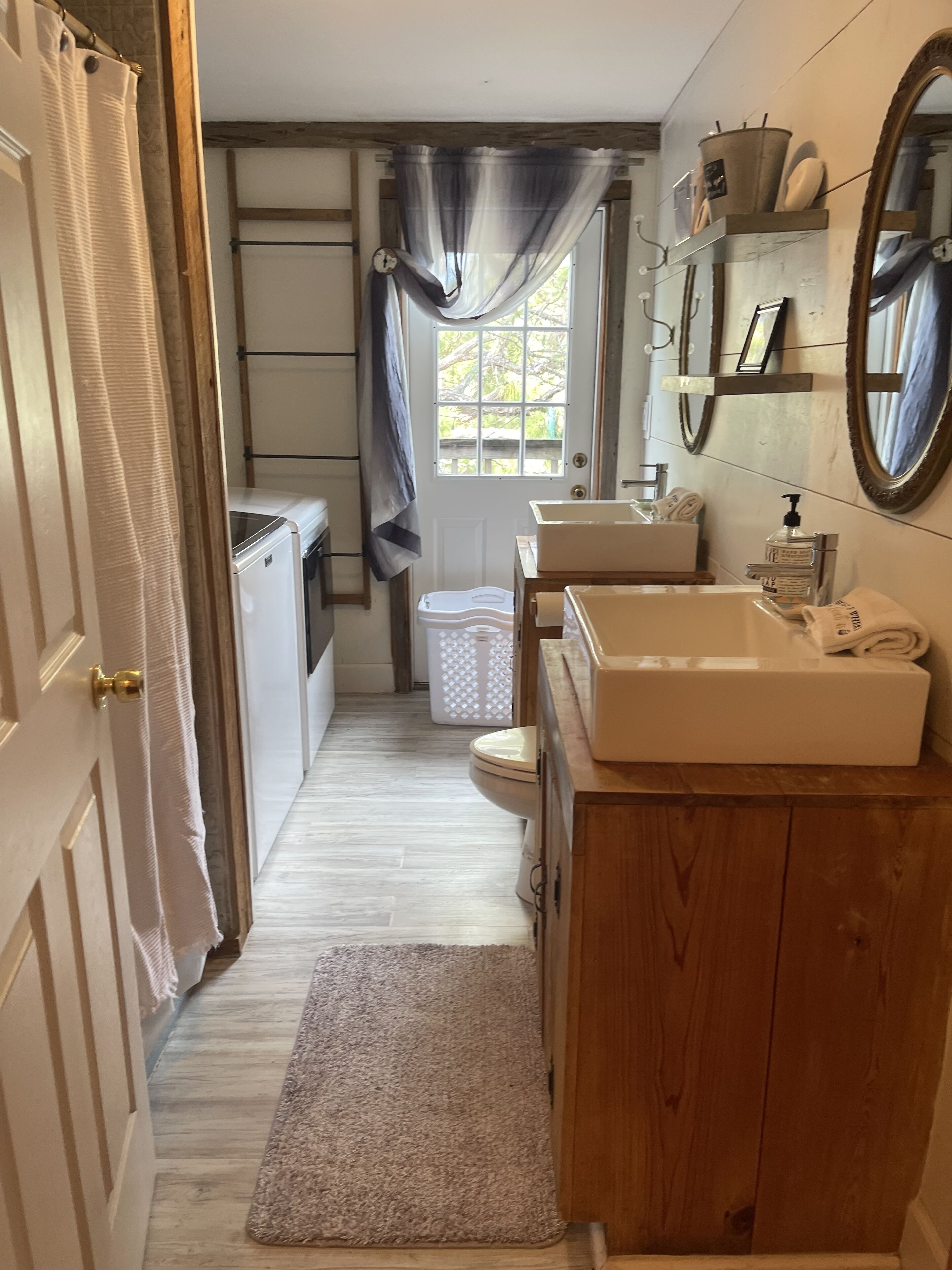 Een kleine badkamer met een houten wastafel, een witte wasbak en een toilet. Er is een raam met gordijnen, een spiegel en een plank met toiletartikelen.