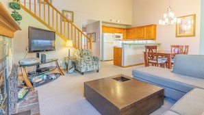 Ein Wohnzimmer mit Sofa, Couchtisch, Fernseher und einer Treppe, die zu einer Küche mit Holzausstattung und einem Essbereich führt.