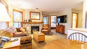 A living room with a fireplace, a television, a dining table, and a comfortable seating area. - Aspens #209 Prime Ski-in Ski-out Location! Pool, Hot tubs, Ski Hill View (Whistler)