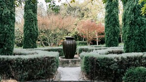 Un jardín formal con un gran jarrón central, setos recortados y árboles altos.