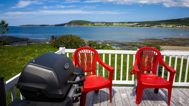 Une terrasse avec un barbecue et deux chaises rouges, donnant sur une étendue d’eau et sur des collines lointaines.