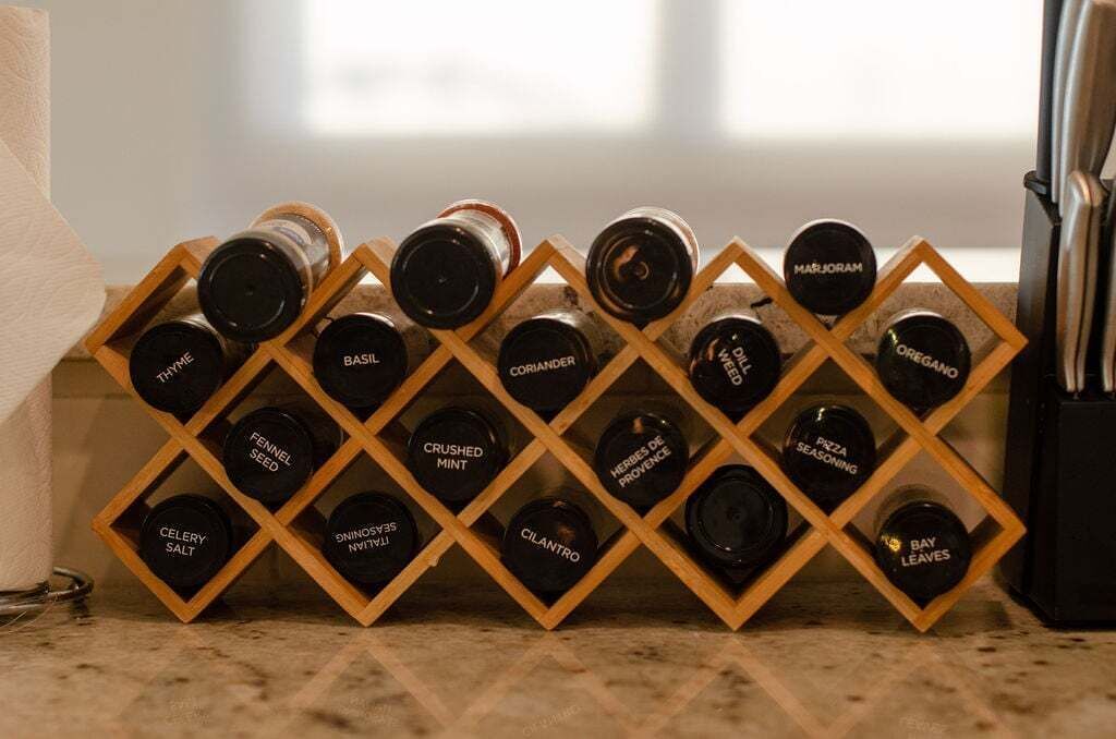 A kitchen counter with a wooden spice rack holding various bottles labeled with names like 'CELERY SALT', 'CRUSHED MINT', and 'BASIL'.