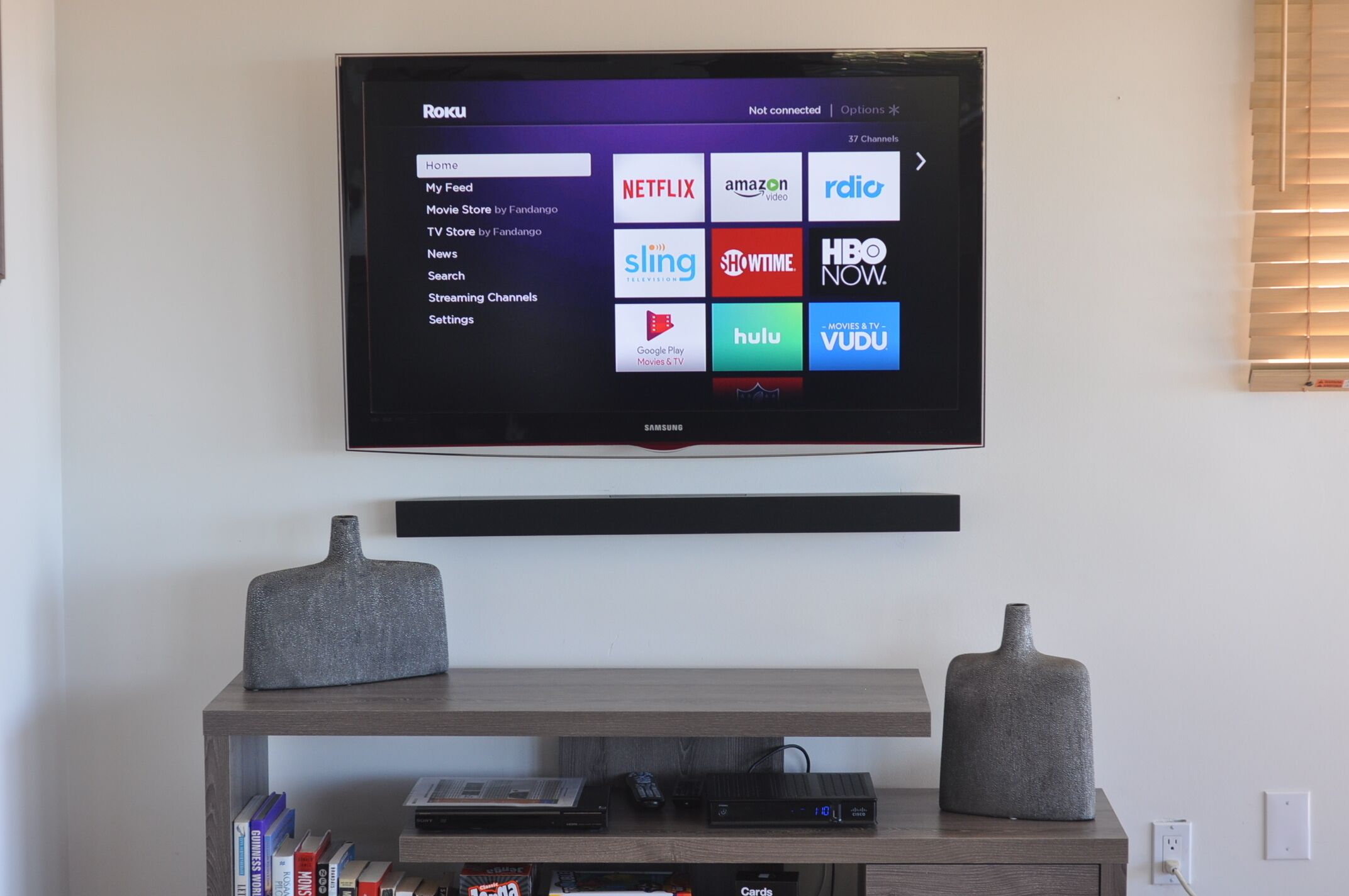 Un televisor montado en la pared muestra una interfaz de Roku con varios iconos de servicios de transmisión.